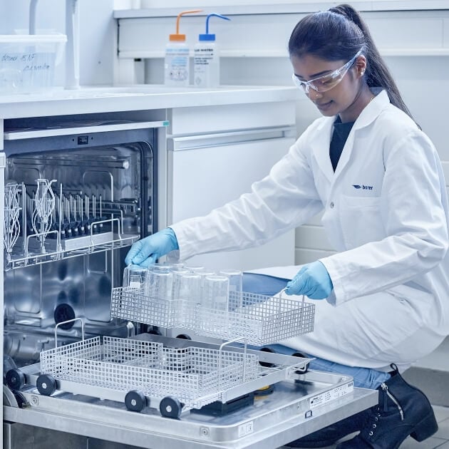 Chemical laboratory technician in protective clothing filling a washing machine with laboratory glassware.